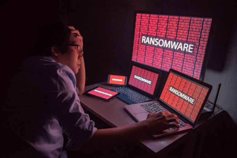 A man at a desk with three laptops and a screen displaying the word "ransomware," emphasizing ransomware protection in Louisville.